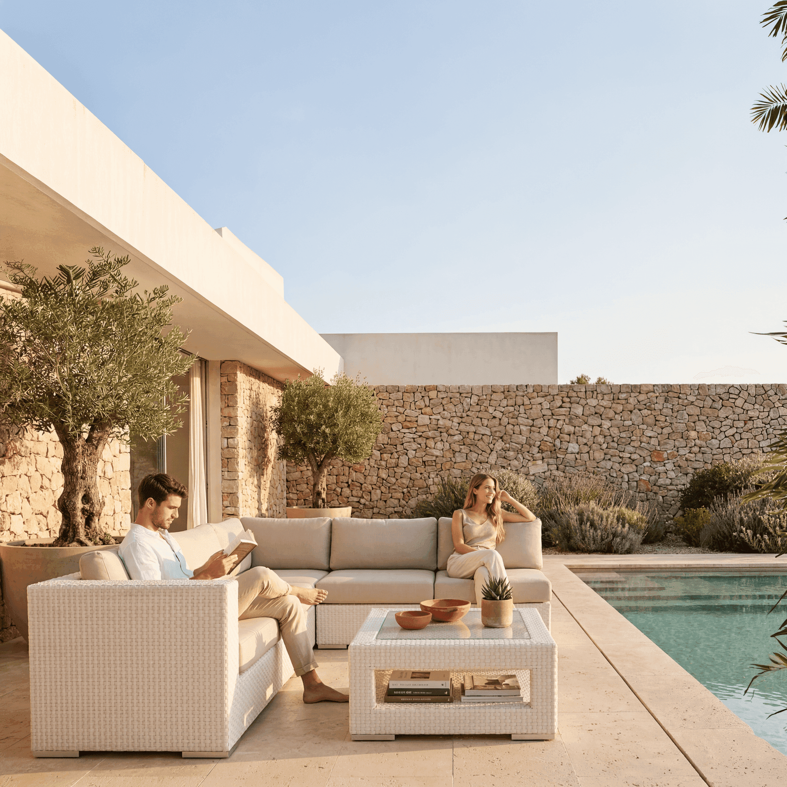Wicker outdoor lounge set with beige cushions and glass coffee table by a pool on a stone terrace at a Mediterranean villa.